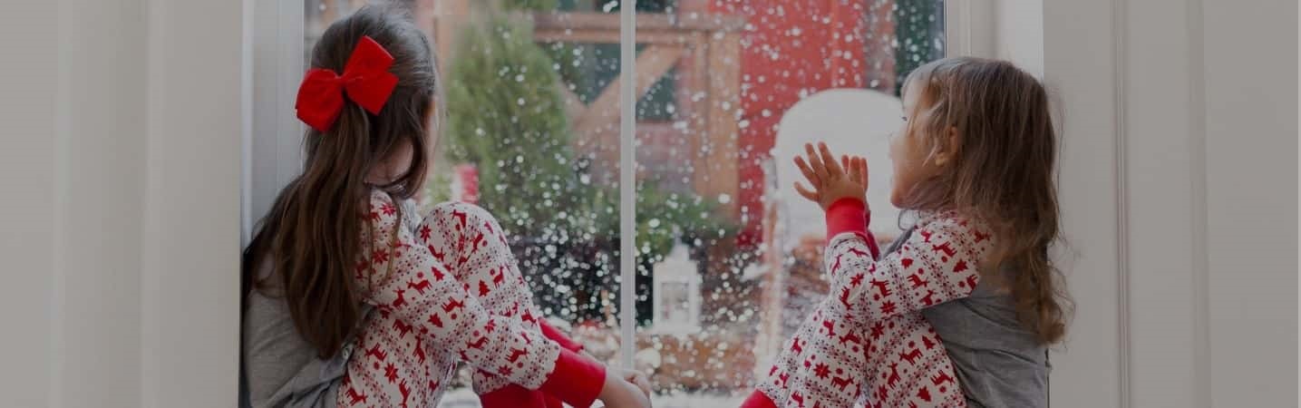 Two cute girls in matching pjs looking out window at snowfall.